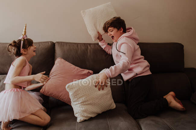 Vista laterale del ragazzo adolescente scalzo che colpisce la sorella in costume da unicorno con cuscino mentre gioca sul divano insieme — Foto stock