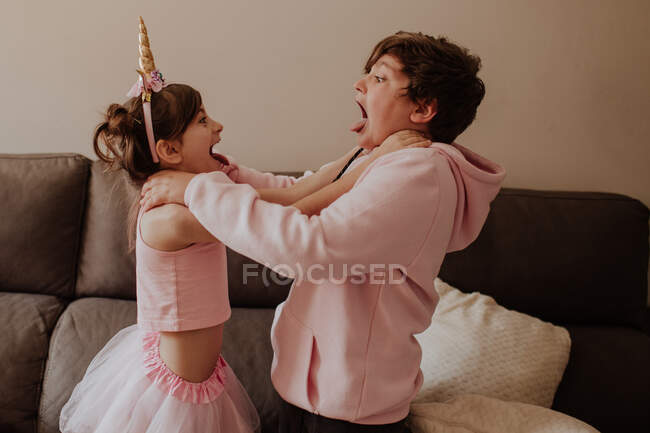 Side view of brother and sister in pink outfits strangle each other while fighting near sofa at home — Stock Photo