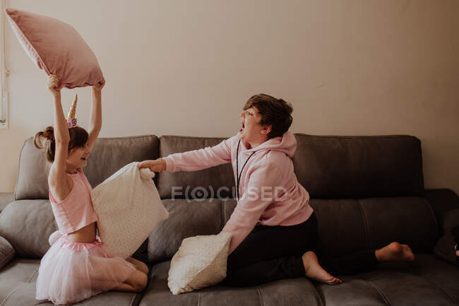 Бічний вид на босоногого підлітка, який б'є сестру в костюмі єдинорога з подушкою під час гри на дивані разом — стокове фото