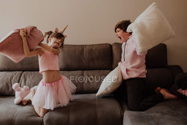 Vista lateral de chico adolescente descalzo golpeando a hermana en traje de unicornio con almohada mientras juegan en el sofá juntos - foto de stock