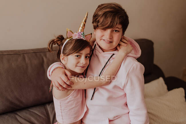 Adolescente chico abrazando lindo hermana mientras de pie cerca cómodo sofá en acogedor habitación en casa - foto de stock