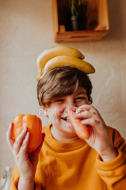 Gordito adolescente chico con plátanos en la cabeza sonriendo y jugando con pimienta y zanahoria en la cocina - foto de stock