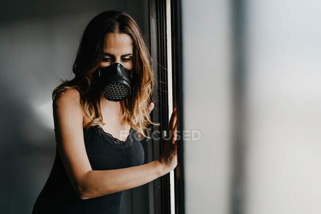 Нещаслива молода жінка в чорній захисній масці, що стоїть біля скляної стіни в закритій кімнаті, представляючи концепцію обмеження та ізоляції під час спалаху коронавірусу — стокове фото