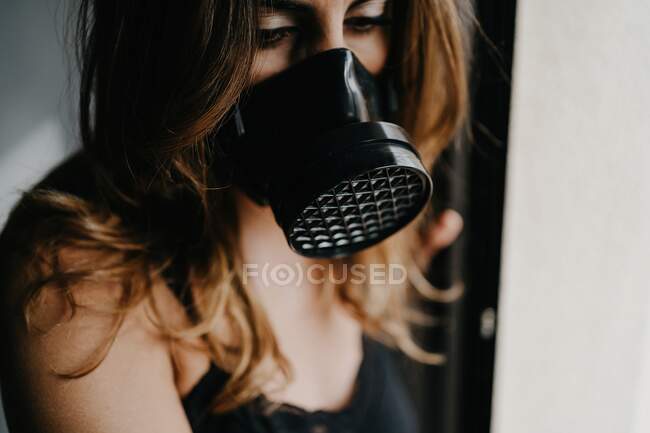 Jeune femme malheureuse portant un masque de protection noir debout près d'une paroi de verre dans une pièce fermée, tout en représentant le concept de restriction et d'isolement pendant l'éclosion de coronavirus — Photo de stock