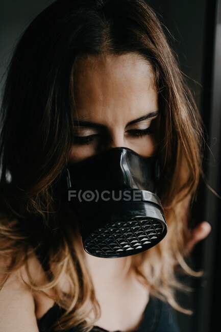 Mujer joven infeliz con máscara protectora negra de pie cerca de la pared de vidrio en una habitación cerrada mientras representa el concepto de restricción y aislamiento durante el brote de coronavirus - foto de stock