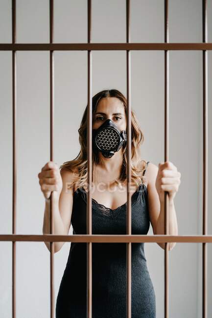 Mujer joven con máscara respiratoria protectora y vestido negro detrás de una valla metálica y mirando a la cámara mientras representa el concepto de prevención y aislamiento del coronavirus - foto de stock