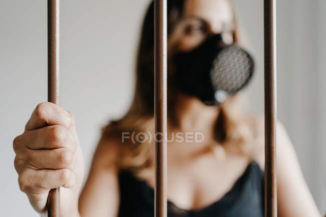 Jeune femme en masque respiratoire protecteur et robe noire debout derrière une clôture métallique et regardant loin tout en représentant le concept de prévention et d'isolement du coronavirus — Photo de stock
