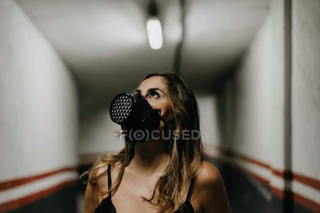 Cheerful young female in elegant black dress and black respirator mask looking up while standing in narrow corridor inside building — Stock Photo