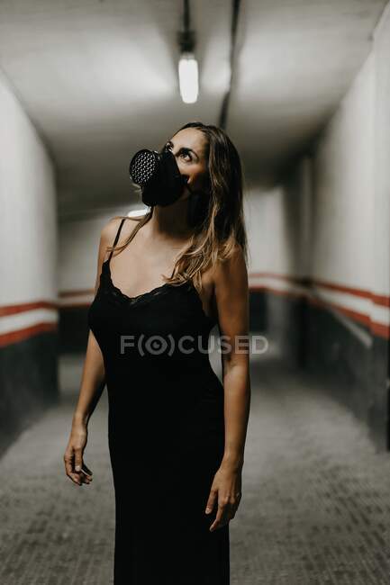 Joven y alegre hembra en elegante vestido negro y máscara respiradora negra mirando hacia arriba mientras está de pie en el pasillo estrecho dentro del edificio - foto de stock