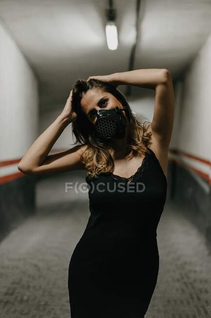 Cheerful young female in elegant black dress and black respirator mask looking at camera while standing in narrow corridor inside building — Stock Photo