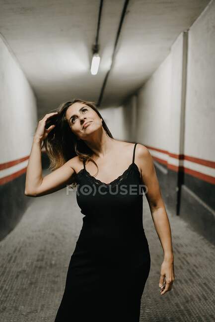 Jovem fêmea em elegante vestido preto de pé em passagem subterrânea fechada e olhando para cima sonhadoramente — Fotografia de Stock