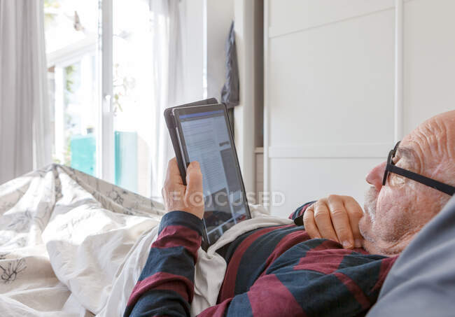Vista lateral do homem idoso em óculos deitado na cama e ler revista eletrônica no tablet enquanto passa a manhã em casa — Fotografia de Stock