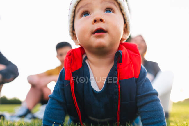 Милий етнічний малюк у теплому одязі посміхається під час повзання на зеленому лузі під час сімейних вихідних у сільській місцевості — стокове фото