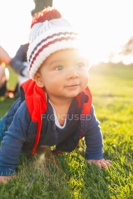 Nettes ethnisches Kleinkind in warmer Kleidung lächelt beim Krabbeln auf der grünen Wiese während des Familienwochenendes in der Natur — Stockfoto