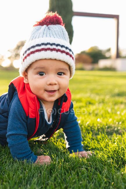 Cute ethnic toddler in warm clothes smiling and looking at camera while crawling on green meadow during family weekend in countryside — Stock Photo
