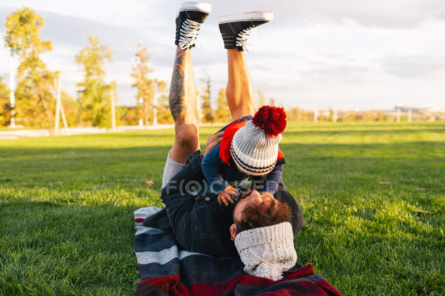 Вид сбоку на веселого молодого человека, лежащего на одеяле на зеленой траве и держащего милого малыша в теплой одежде во время отдыха на выходных в парке — стоковое фото