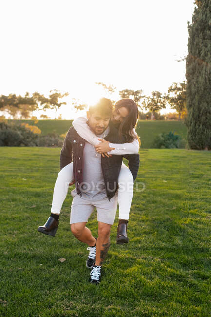 Jovem alegre dando passeio de piggyback para a esposa feliz enquanto desfruta do dia ensolarado de outono no prado verde no campo — Fotografia de Stock