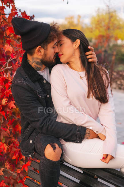 Felice giovane uomo e donna abbracciare e baciare mentre seduto sulla panchina vicino colorato albero autunnale nel parco — Foto stock