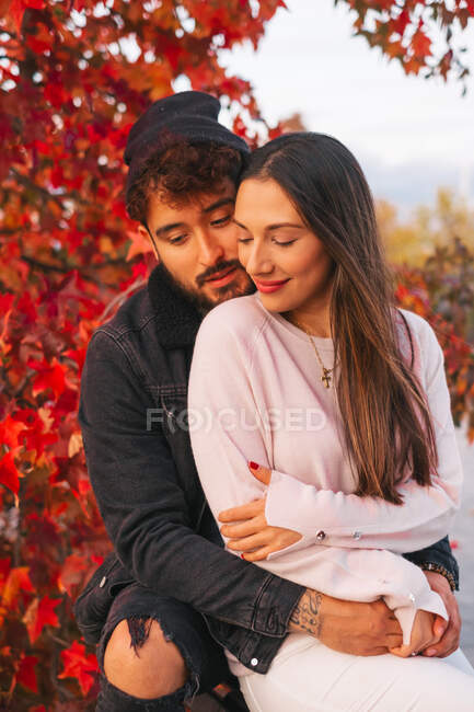 Feliz joven hombre y mujer abrazando mientras está sentado en el banco cerca de colorido árbol de otoño en el parque - foto de stock