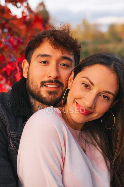 Glückliche junge Mann und Frau umarmen sich, während sie auf Bank in der Nähe von bunten Herbstbaum im Park sitzen — Stockfoto