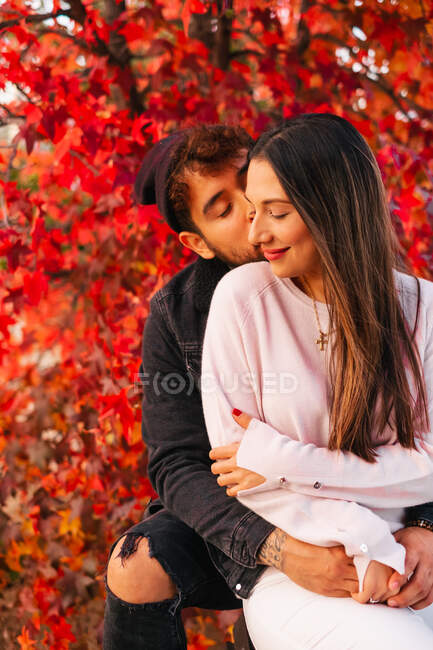 Glückliche junge Mann und Frau umarmen und küssen sich, während sie auf Bank in der Nähe von bunten Herbstbaum im Park sitzen — Stockfoto