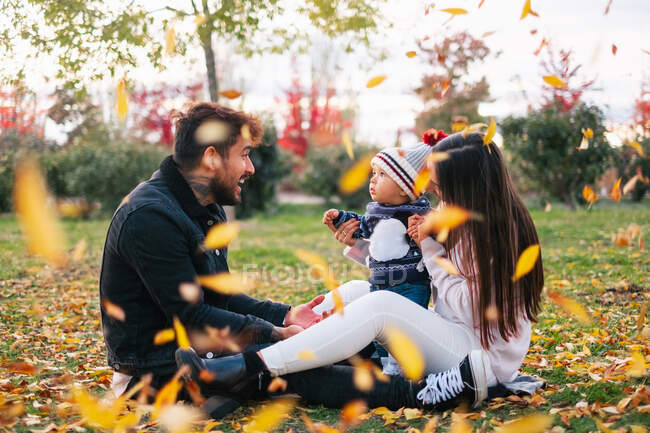 Side view of cheerful young man and woman with cute little baby sitting on grass and having fun with yellow leaves while spending time together in autumn park — Stock Photo