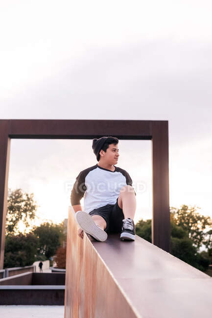 Niedriger Winkel positiver junger Mann in Freizeitkleidung sitzt auf einem Steinzaun und schaut weg, während er sich im Stadtpark bei sonnigem Abend ausruht — Stockfoto