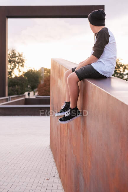 Baixo ângulo de homem jovem positivo no desgaste casual sentado na cerca de pedra e olhando para longe enquanto descansa no parque da cidade na noite ensolarada — Fotografia de Stock