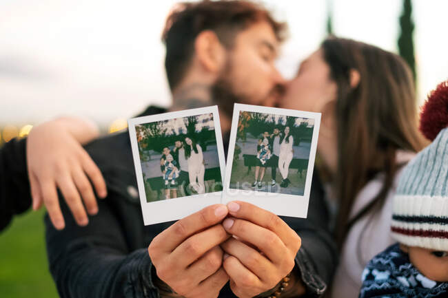 Счастливая молодая супружеская пара показывает мгновенные фотографии семьи, стоя с детьми и целуясь в парке — стоковое фото