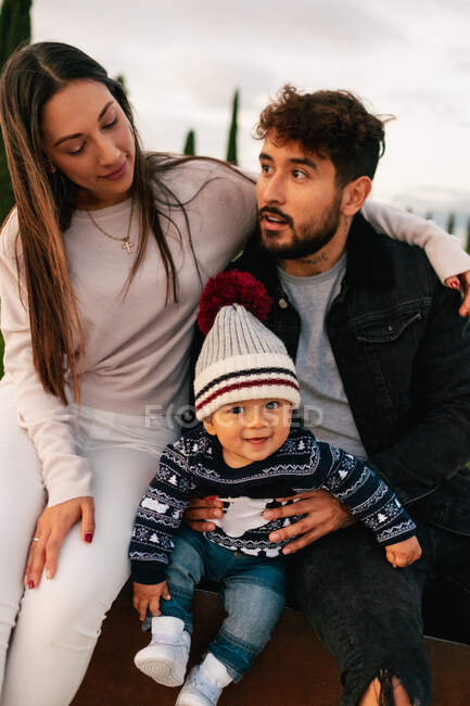 Positive young parents with cute toddler kid in casual warm clothes embracing while sitting together on street in cloudy autumn day — Stock Photo