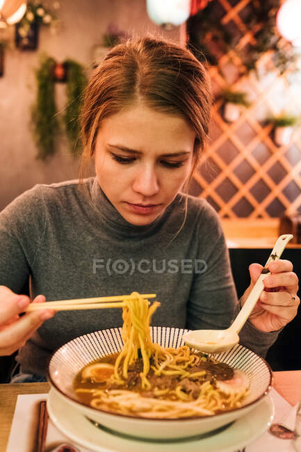 Jovem fêmea usando pauzinhos e colher para comer ramen saboroso enquanto se senta à mesa no restaurante japonês — Fotografia de Stock
