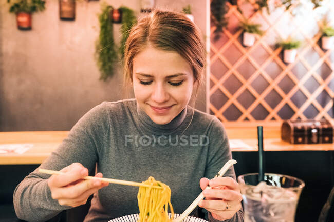 Jeune femme utilisant des baguettes et une cuillère pour manger de savoureux ramen assis à table dans un restaurant japonais — Photo de stock