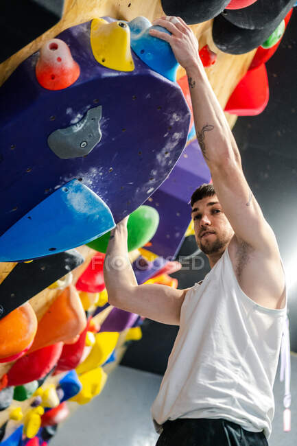 From below of young man in sportswear hanging on climbing wall and looking at camera during bouldering workout in gym — Stock Photo