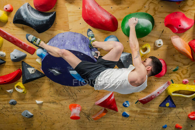 Вид сбоку сильного мужчины в спортивной одежде, высоко взбирающегося на красочные захваты стены во время тяжелой тренировки в тренажерном зале — стоковое фото