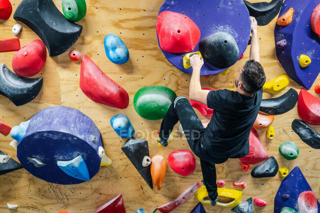 Обратный вид на неузнаваемого сильного спортсмена в спортивной одежде, лазающего по красочной стене во время тренировки в современном альпинистском центре — стоковое фото