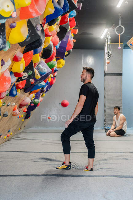 Вид сбоку на вдумчивого молодого человека в спортивной одежде, стоящего и смотрящего на скалолазную стену с размытым мужчиной, сидящим на заднем плане — стоковое фото