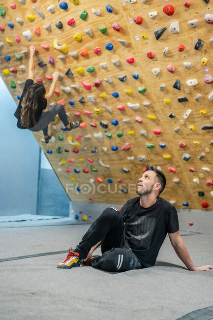 Sportlicher Mann in Sportbekleidung sitzt auf dem Boden und schaut nach oben, während unscharfe anonyme Bergsteigerin an der Wand im Fitnessstudio trainiert — Stockfoto
