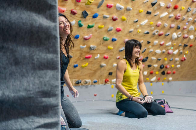 Happy female athletes sitting in lotus on knees on the floor while smiling and looking away resting during climbing training in gym — Stock Photo