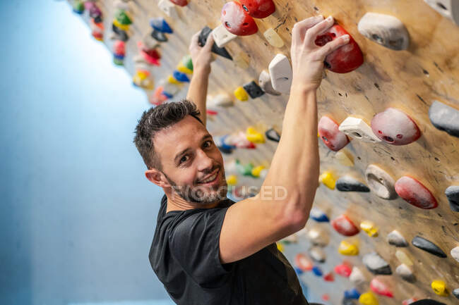 Seitenansicht eines starken, glücklichen männlichen Athleten, der während des Trainings in einem modernen Fitnessstudio in Sportbekleidung auf eine bunte Wand klettert — Stockfoto