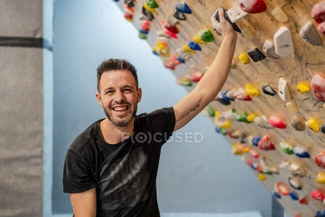 Joyful young male climber in sporty wear laughing and looking at camera with hand on climbing wall in modern gym — Stock Photo