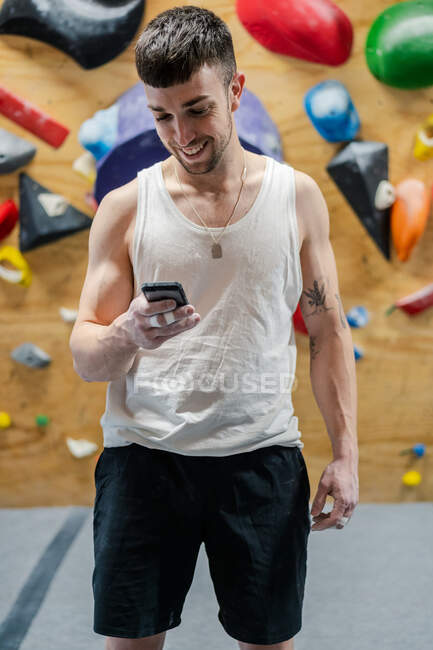 Giovane sportivo muscolare in piedi usura attiva e utilizzando il telefono cellulare durante l'allenamento di boulder in palestra — Foto stock