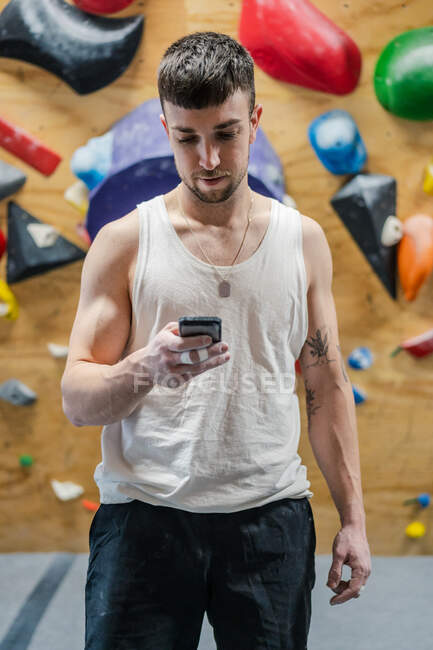 Junger muskulöser Sportler in aktiver Kleidung im Stehen und mit Handy beim Bouldertraining im Fitnessstudio — Stockfoto
