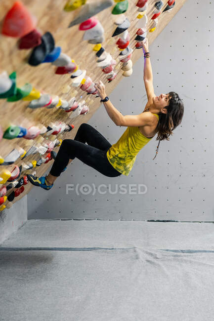 Seitenansicht einer fokussierten Frau in Sportbekleidung, die an einer steilen Wand über Matten im modernen Kletterzentrum hängt — Stockfoto