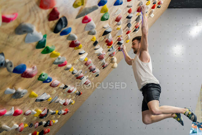 Vista lateral desde abajo del hombre joven en desgaste activo sosteniendo apretones firmemente mientras cuelga en el aire durante el entrenamiento de escalada en el gimnasio - foto de stock