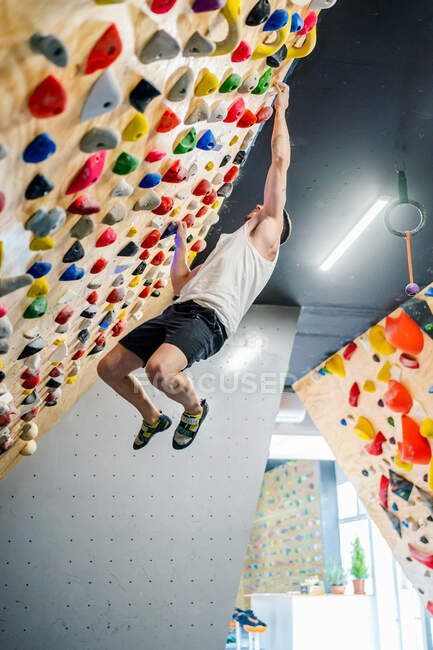 Vue latérale d'en bas du jeune homme en tenue active tenant fermement les poignées tout en restant suspendu dans l'air pendant l'entraînement d'escalade dans la salle de gym — Photo de stock