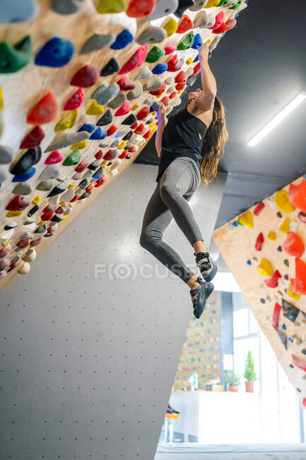 Vista lateral de baixo de mulher focada em sportswear pendurado na parede íngreme acima tapetes no moderno centro de escalada — Fotografia de Stock