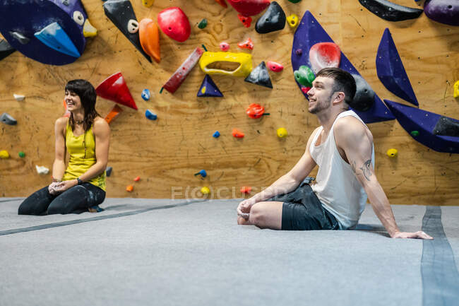 Вид сбоку улыбающихся мужчины и женщины, сидящих на полу и улыбающихся во время тренировки в скалодроме — стоковое фото