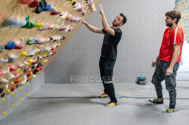 Vista lateral do atleta masculino em sportswear escalada em apertos de parede íngreme, enquanto amigo masculino apoiar com saco de talco no ginásio — Fotografia de Stock