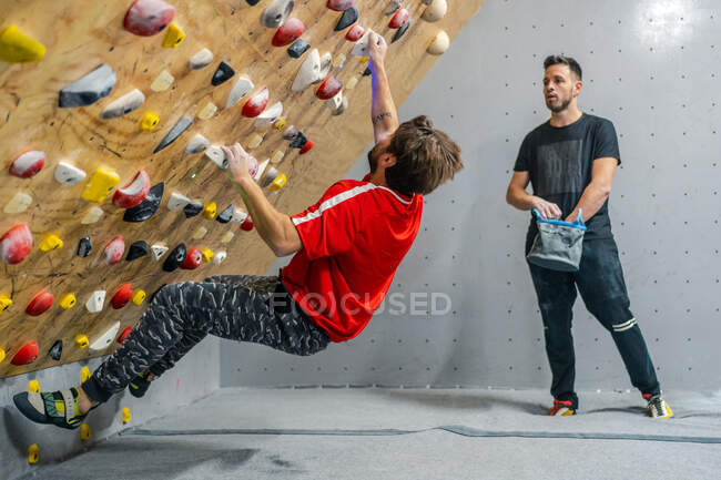Side view of male athlete in sportswear climbing on grips of steep wall while male friend supporting with bag of talcum in gym — Stock Photo