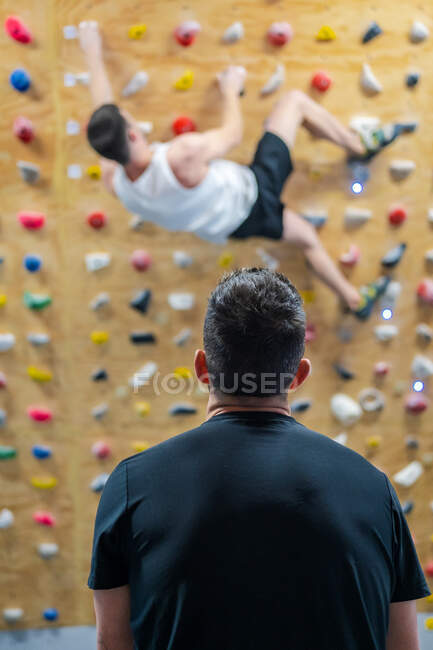 Вид ззаду анонімного спортсмена-чоловіка в спортивному одязі, який спостерігає за альпіністом під час тренувань на стіні в спортзалі — стокове фото